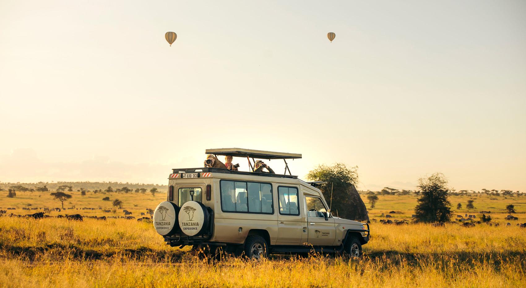 Ein Tanzania-Experience-Fahrzeug in der Serengeti. Zwei Heißluftballons sind am Himmel zu sehen. Ein Tanzania-Experience-Fahrzeug in der Serengeti. Zwei Heißluftballons sind am Himmel zu sehen.
