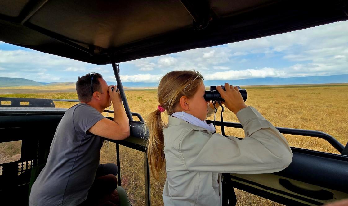 A father and his daughter are in a safari vehicle, gazing through the roof hatch into the savannah with binoculars.