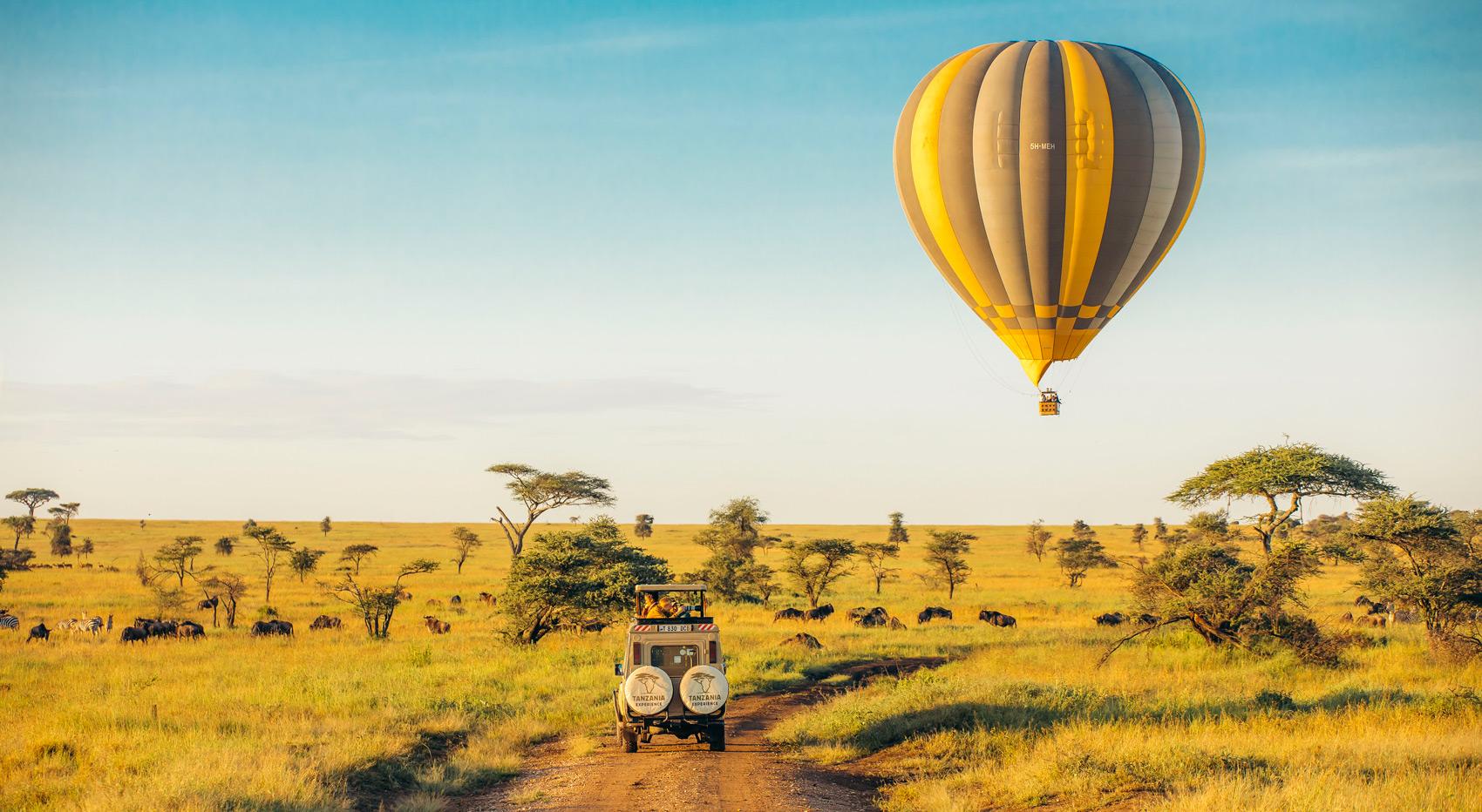 Ein Heißluftballon schwebt elegant über der Serengeti, während ein Tanzania-Experience Safarifahrzeug darunter unterwegs ist. Ein Heißluftballon schwebt elegant über der Serengeti, während ein Tanzania-Experience Safarifahrzeug darunter unterwegs ist.