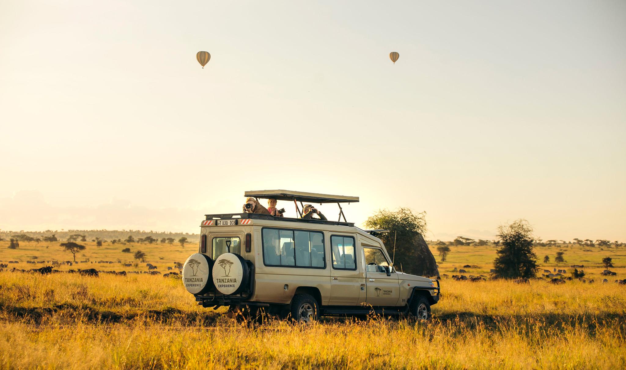 Ein Safarifahrzeug von Tanzania-Experience auf einer Pirschfahrt in Tansania mit Heißluftballons im Hintergrund.