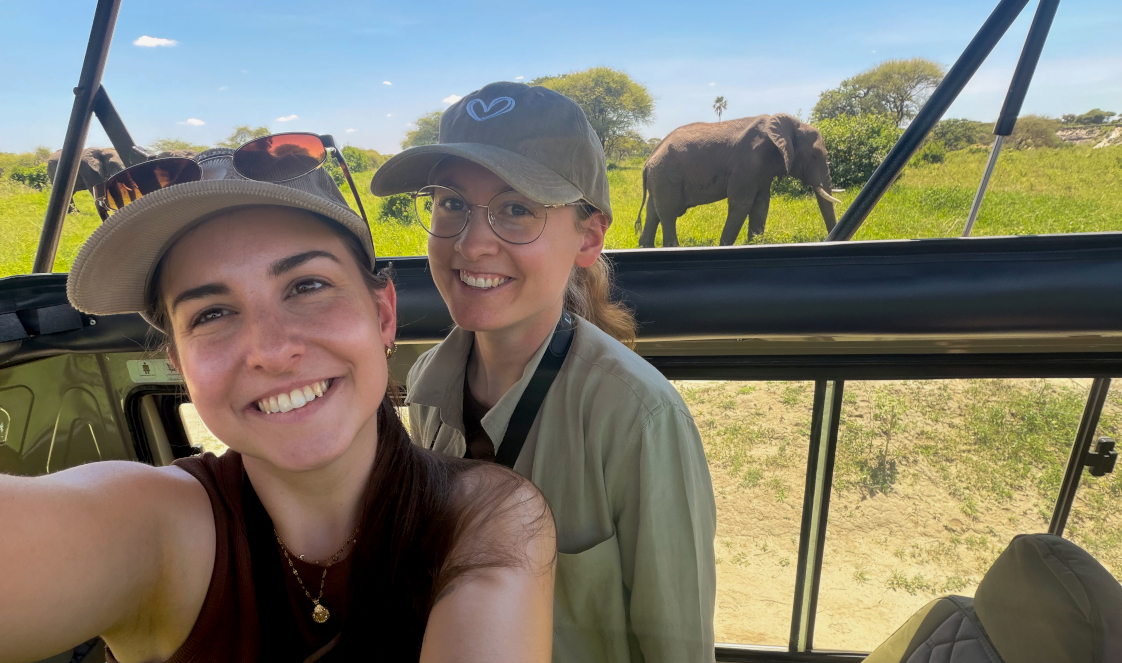 Two guests with Tanzania-Experience stand in the roof hatch of their safari vehicle, taking a selfie with a small herd of elephants in the background.