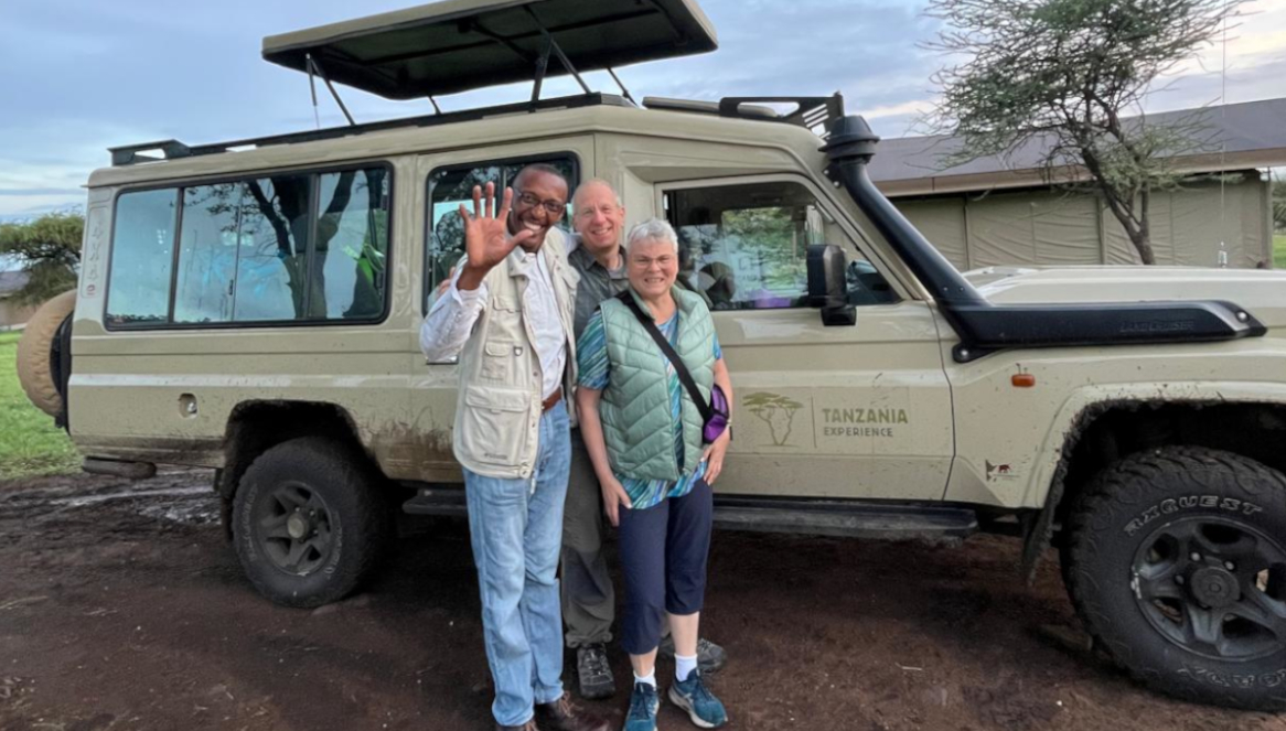 Alfred, the Tanzania-Experience guide, is standing with his two guests in front of their safari vehicle.
